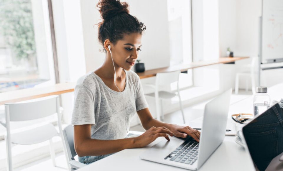 Romantic african girl with trendy hairstyle sitting at her workplace and analysing data. Indoor portrait of black female student working with laptop before exam..